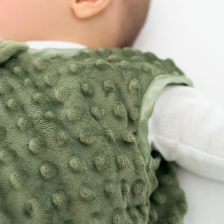 A baby lies wrapped in the HALO Wearable Blanket Plushy Dot Velboa TOG 1.5, a soft green blanket with raised dots, over a white long-sleeve shirt. Part of the baby's face shows, highlighting cozy comfort and safe sleep.