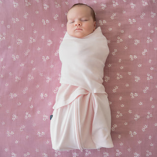A baby sleeps wrapped snugly in the HALO Swaddle 100% Organic Cotton TOG 1.5, lying on a pink blanket with small white floral patterns.