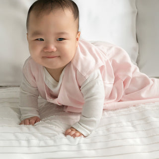 A smiling baby crawls on a white bedspread, dressed in the HALO Wearable Blanket 100% Organic Cotton TOG 0.5—a light pink, sleeveless sleep sack over a white long-sleeve outfit—ready for safe and cozy sleep.