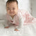 A smiling baby crawls on a white bedspread, dressed in the HALO Wearable Blanket 100% Organic Cotton TOG 0.5—a light pink, sleeveless sleep sack over a white long-sleeve outfit—ready for safe and cozy sleep.