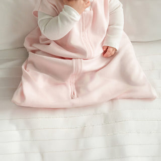 A baby in the HALO Wearable Blanket 100% Organic Cotton TOG 0.5 and a white long-sleeve shirt sits on a white bed, hand near their mouth, enjoying a cozy and safe sleep.