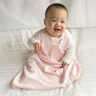 A smiling baby in a HALO Wearable Blanket 100% Organic Cotton TOG 0.5 and a white long-sleeve shirt sits on a textured white blanket, ready for safe sleep.