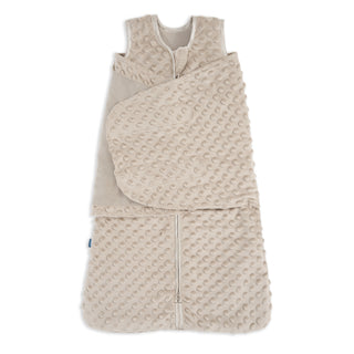 The HALO Swaddle Plushy Dot Velboa TOG 3.0 is a beige, sleeveless sleep sack with bumpy fabric, front zipper, and an attached adjustable swaddle wrap for cozy comfort.