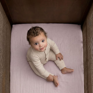 A baby with curly hair, in a light beige outfit, sits barefoot on HALO Fitted Crib Sheets 100% Organic Cotton Muslin 2 Pack in a wooden crib, gazing up at the camera with curiosity.