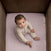A baby with curly hair, in a light beige outfit, sits barefoot on HALO Fitted Crib Sheets 100% Organic Cotton Muslin 2 Pack in a wooden crib, gazing up at the camera with curiosity.