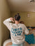 A woman with her hair in a bun faces away, holding a baby and giving a thumbs down. She wears the HALO Tired Moms Club Sweatshirt in bold blue letters. The room features a bed with clothes scattered and light green walls.