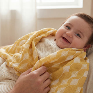 A smiling baby lies on their back, wrapped in an aden + anais essentials cotton muslin swaddle blanket with yellow and white patterns, gently held by an adult. The baby wears a cream outfit indoors with soft natural light.