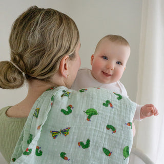 A woman with blonde hair in a low bun holds a smiling baby over her shoulder. Draped over her shoulder is an aden + anais essentials cotton muslin swaddle blanket featuring caterpillar and butterfly prints.