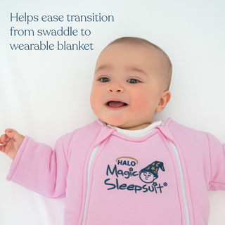 A smiling baby lies on a white surface, wearing a pink HALO Baby Merlin's Magic Sleepsuit® 100% Cotton. Blue text reads, Helps ease transition from swaddle to wearable blanket—a cozy innovation in baby sleepwear.
