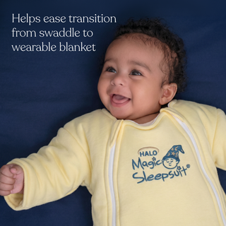 A smiling baby lies on a dark blue sheet in a yellow Baby Merlin’s Magic Sleepsuit® 100% Cotton by HALO. White text: Eases the transition from swaddle to wearable blanket—perfect for smooth swaddle transitions and restful nights.
