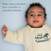 A smiling baby lies on a light blue surface wearing the HALO Baby Merlin's Magic Sleepsuit® 100% Cotton. Text beside the baby reads, Helps ease transition from swaddle to wearable blanket for a smoother swaddle transition.