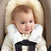 A baby with short brown hair sits in a car seat, wearing a cream sweater and aden + anais essentials cotton muslin strap covers featuring elephant prints. The seat has a soft beige interior and a black strap across the chest. The smiling baby glances slightly to the side.