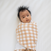 A dark-haired baby is wrapped in a beige and white gingham HALO Swaddle Micro-Fleece TOG 3.0, lying on a white surface. The award-winning design ensures healthy hip development, and the baby looks content.