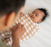 A parent swaddles a baby in HALOs Swaddle Micro-Fleece TOG 3.0, renowned for promoting healthy hip development. The cozy, checkered wrap features beige and white squares, keeping the baby content and relaxed on a white surface.