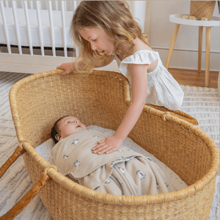 A young girl with curly hair, in a white dress, gently places her hand on a baby wrapped in a HALO Swaddle Micro-Fleece TOG 3.0, lying in a wicker bassinet. The room features a crib, small table, and patterned rug—ideal for healthy hip development with its adjustable wings.