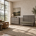 A gray Child Craft Surrey Hill 2-Piece Nursery Set with a 4-in-1 convertible crib and six-drawer dresser in a nursery with a leather armchair and a large window.