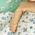 A baby in a beige sweater is lying down, partially covered by a patterned blanket with dinosaurs, the moon, and playful designs. Beside them is a green swaddle from the Disney™ Essentials cotton muslin swaddle blanket 4 pack by aden + anais featuring similar cartoon motifs.