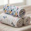 Two rolled-up aden + anais essentials cotton muslin swaddle blankets, one with small car illustrations and the other with a bright checkered pattern, rest on a beige surface. A softly blurred window is visible in the background.