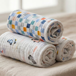 Two rolled-up aden + anais essentials cotton muslin swaddle blankets, one with small car illustrations and the other with a bright checkered pattern, rest on a beige surface. A softly blurred window is visible in the background.
