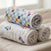 Two rolled-up aden + anais essentials cotton muslin swaddle blankets, one with small car illustrations and the other with a bright checkered pattern, rest on a beige surface. A softly blurred window is visible in the background.