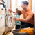 In a snug laundry room, a man in orange helps a toddler in a blue shirt operate the washing machine. Beside them, clothes sit in a basket with HALOs DreamWeave Breathable Crib Cover, ensuring comfort for the little one as he watches attentively.