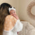 A woman with long hair cradles a baby on her shoulder, who wears a white headband with a bow. The baby rests comfortably against an absorbent aden + anais boutique 100% cotton muslin burpy bib, while behind them a decorative mirror enhances the warmth of the scene.
