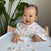 A smiling baby in a high chair reaches for snacks on the tray, wearing HALOs cheerful Tried & True Bundle Burpy Bib with rainbows. Large green leaves in the background add a natural touch to this joyful scene.