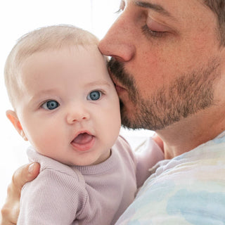 A bearded man gently kisses the cheek of a baby with big blue eyes, wrapped in aden + anais organic cotton muslin burpy bibs. The baby, dressed in light purple, gazes into the distance with an open mouth expression against a softly lit, beautifully blurred background.