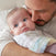 A man gently holds a baby wrapped in an aden + anais organic cotton muslin burpy bib, pastel-colored; the baby gazes curiously to the side as he affectionately nuzzles their cheek against a softly blurred backdrop.