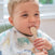 A baby with short blonde hair sits holding a beige spoon, its tip in their mouth. Theyre wearing an aden + anais boutique silky soft burpy bib featuring colorful animal prints. The background is softly blurred.