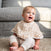 A happy baby with light hair and blue eyes sits on a textured carpet, wearing a beige bib from the aden + anais boutique silky soft burpy bib 2 pack. Its perfect for spills and pairs well with the babys light-colored outfit, all set against a gray sofa backdrop.