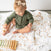 A toddler in a green outfit sits on an aden + anais organic cotton dream blanket, playing with a colorful xylophone. They wear a small bow headband and are focused on their activity.