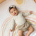A baby smiles while lying on an aden + anais boutique cotton muslin dream blanket with sun and rainbow patterns, wearing a light-colored outfit and a headband with a bow for extra charm and comfort.