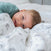 A smiling blond toddler lies on a bed, partially covered by an aden + anais organic cotton dream blanket with gray animal prints. Wearing a teal shirt and looking toward the camera, the child creates a playful and cozy atmosphere.