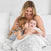 A woman smiles on a bed, holding a baby wrapped in an aden + anais boutique silky soft muslin dream blanket, while a young girl kisses the babys cheek in a bright, cozy setting with white pillows and bamboo-infused viscose bedding.