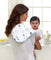 A woman holding a baby, looking at the camera, has an aden + anais boutique 100% cotton muslin burpy bib with star patterns draped over her shoulder. The softly lit window highlights its design, while the counter displays various kitchen items.