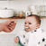A smiling baby wears an aden + anais boutique 100% cotton muslin burpy bib, adorned with stars, while being fed by an adult. The absorbent design ensures a mess-free mealtime. In the background, kitchen shelves display dishes, jars, and cups.