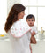 A woman with long brown hair, in a white shirt, cradles a baby dressed in white. Over her shoulder is an aden + anais boutique 100% cotton muslin burpy bib featuring heart patterns. They are in a bright room with large windows behind them.