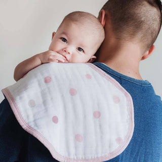 A baby rests on a persons shoulder, looking at the camera. The person wears a blue shirt with an aden + anais boutique 100% cotton muslin burpy bib featuring pink polka dots. The softly blurred background adds a luxurious finish to this heartwarming moment.