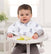 A baby wearing an aden + anais boutique 100% cotton muslin burpy bib sits in a highchair with green apple slices on the tray. The bright room features light-colored decor and a small plant in the background.