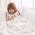 A smiling young girl with long hair sits on a white bed wrapped in an aden + anais boutique cotton muslin dream blanket, offering breathable comfort. The 100% cotton muslin features pink and beige heart patterns, while bright light filters through the window behind her.
