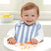 A smiling light brown-haired baby sits in a high chair wearing an aden + anais blue and white 100% cotton muslin snap bib from the 3-pack. The absorbent bib complements the babys grip on an orange spoon with cereal pieces, while a bowl of lemons blurs in the background.