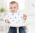 A baby in a high chair wears an aden + anais boutique 100% cotton muslin snap bib featuring a bird pattern. The adjustable, absorbent bib matches the babys cream sweater and blue jeans, set against a bright kitchen backdrop.