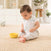 A baby, wearing aden + anais boutique cotton muslin snap bibs, sits on a beige textured rug, entranced by a yellow bowl. The cozy setting includes light clothing with small designs, a plant, and white cabinets.