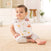 A baby sits on a beige carpet, wearing an aden + anais Boutique 100% cotton muslin snap bib with colorful illustrations. With short brown hair, the baby sports a white shirt and light gray patterned pants. A white cabinet and plant enhance the cozy background.