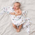 A smiling baby is wrapped in a breathable, colorful aden + anais boutique 100% cotton muslin swaddle blanket, lying on a cream-colored surface.