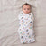A baby on a gray bed gazes upwards, wrapped in an aden + anais boutique 100% cotton muslin swaddle blanket adorned with colorful planets and rockets, with a tuft of dark hair peeking out.