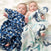 Two babies lie on a soft, white surface wrapped in aden + anais boutique 100% cotton muslin swaddles. The left baby is in a blue polka dot, while the right one is in a breathable white blanket with blue and teal patterns.