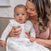 A woman lovingly holds a baby in a white outfit with light blue patterns, wrapped in an aden + anais organic cotton light sleep sack TOG 1.0. They sit on a couch, the woman smiling and gently resting her head next to the baby, creating a warm, affectionate atmosphere.