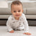 A baby in a white onesie with gray patterns crawls on a light carpet beside an aden + anais organic cotton light sleep sack TOG 1.0. The baby has curly hair, a curious expression, and the cozy sleep sack with a gray sofa in the background.